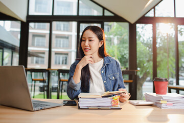 Smiling young Asian woman clearing paperwork in office, using laptop and many documents beside her. Accounting and finance business concept.