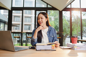 Smiling young Asian woman clearing paperwork in office, using laptop and many documents beside her. Accounting and finance business concept.