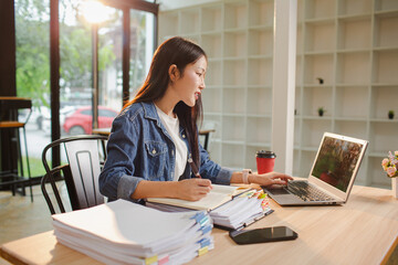 Smiling young Asian woman clearing paperwork in office, using laptop and many documents beside her. Accounting and finance business concept.
