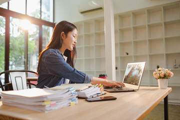 Smiling young Asian woman clearing paperwork in office, using laptop and many documents beside her. Accounting and finance business concept.