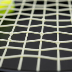 Close-up of a tennis racket's strings, showing the woven pattern and a blurred tennis ball in the background.
