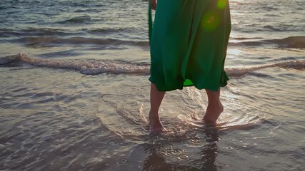 Golden hour sunlight illuminates a woman in a long green dress enjoying a barefoot stroll along the wet sand of a tranquil beach as gentle waves lap at her feet