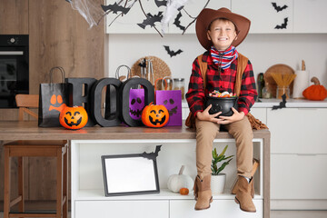 Little boy dressed as cowboy for Halloween with candies on table in kitchen