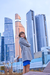 A woman in a blue dress stands in front of a tall building. The unbuttoned coat highlights her slender body.