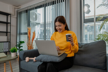 Young Asian woman in yellow sweatshirt smiling while watching something on her laptop and holding a cup of coffee. She sits comfortably on a sofa in a cozy, bright living room at home.