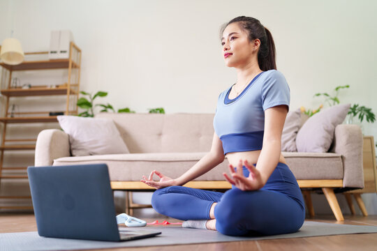 Asian adult woman meditating home practicing yoga online laptop serene pose wellness mindfulness healthy lifestyle indoor exercise fitness