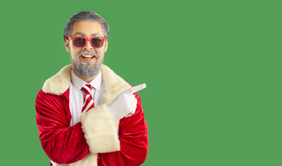 Glad male Santa Claus dressed in red clothing, white gloves and stylish sunglasses, indicating index finger on copy space, showing place for advertisement or promotion, green background