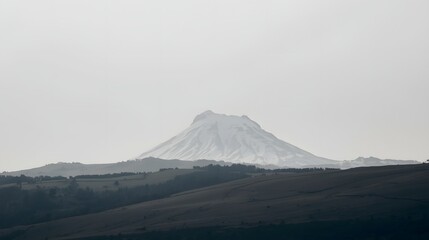Majestic snow-covered mountain peak rising above dark rolling hills under a gray sky, capturing the serene beauty of winter wilderness landscape, perfect for nature background or travel inspiration.