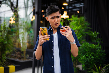 Surprised young Asian man in navy shirt and white t-shirt looks at his declined credit card and smartphone in disbelief. Online payment refused or blocked card.