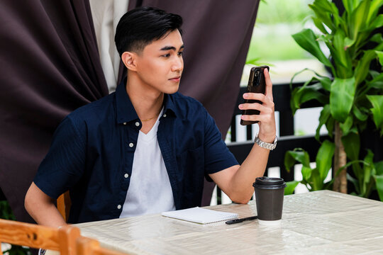 A young Asian man holds his smartphone up while sitting at a garden café with coffee and notebook, as if on a video call or checking updates online.