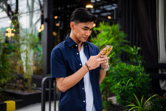 Casual young Asian man in navy shirt and white tee smiles while focused on his smartphone outside a green café garden.