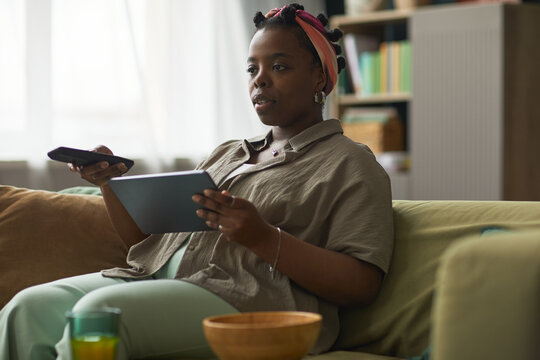 Young adult Black woman sitting on sofa holding digital tablet and remote control, watching television with focused expression, bowl on table in foreground, bookshelf in background