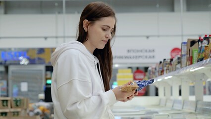 Young woman browsing groceries in a supermarket, carefully reading product details on packaging in the refrigerated section of the store