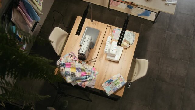No people top view of professional industrial sewing machine and serger on table in workshop, lamps above, patchwork garment at work, rail with handmade merchandise, filmed from upper floor