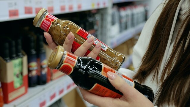 Customer comparing two sauce bottles in a supermarket and making a decision based on ingredients and nutrition facts while grocery shopping