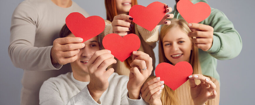 Several young people wishing you happy Valentine's Day. Banner background with group of cheerful, joyful men and women smiling and holding beautiful red heart shaped paper cards in hands. Love concept