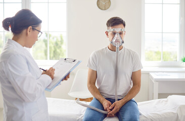 Female doctor with clipboard recording indicators or diagnosis of male patient with breath difficulties, sitting on hospital bed and wearing oxygen mask during rehabilitation or medical treatment.