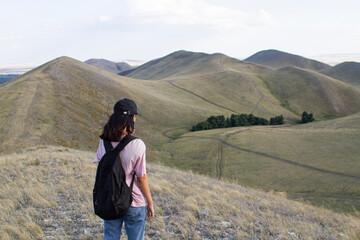 Naklejka premium a teenage girl is standing with her back to the camera, holding a black backpack and looking out at the Karamuruntau mountain range