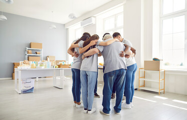 Group of volunteers of different ages indoors standing in a circle hugging, view from the back. Employees of a charity fund before starting work. Concept homeless center, humanitarian project