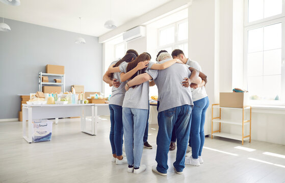 Group of volunteers of different ages indoors standing in a circle hugging, view from the back. Employees of a charity fund before starting work. Concept homeless center, humanitarian project