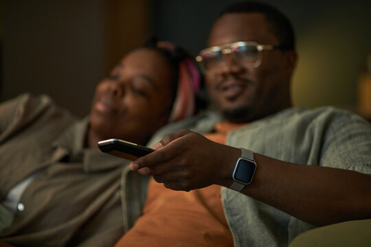 Black man holding remote control sitting next to Black woman relaxing on couch watching television together, both appearing comfortable and engaged in activity - Powered by Adobe