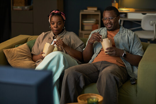 Black woman and Black man sitting on sofa eating takeout food while watching television together, both appearing relaxed and focused on screen, remote control visible on couch - Powered by Adobe