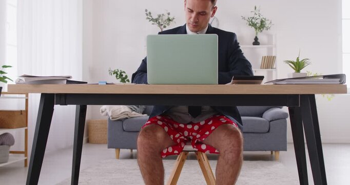 Serious business man wearing a formal suit jacket and funny red polka dot boxer shorts underwear working remotely sitting at a desk with a laptop computer at home