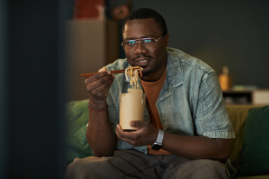 Portrait of young adult Black man wearing glasses sitting indoors eating noodles with chopsticks from takeout container, watching tv with neutral expression - Powered by Adobe