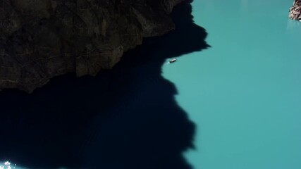 A close-up view of Attabad Lake in Pakistan featuring a small boat on its calm, turquoise waters. The vibrant lake contrasts with steep, rugged cliffs that cast a dark shadow across the surface.