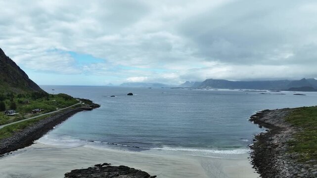 Aerial reverse dolly over Rorvik Beach in Lofoten, Norway revealing Road E10 to Henningsvaer curving around beach on left and right.