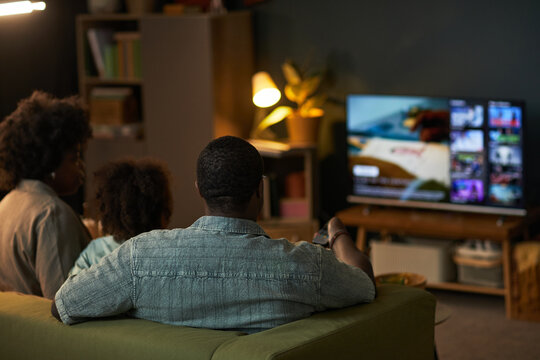 Black man, Black woman, and child sitting on sofa watching television together in living room, remote control in man's hand, family spending leisure time