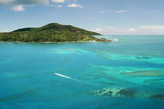Aerial view. The tropical island is surrounded by a beautiful ocean and coral reefs. A speedboat is leaving the white trail on the waves. Inner islands of Seychelles.