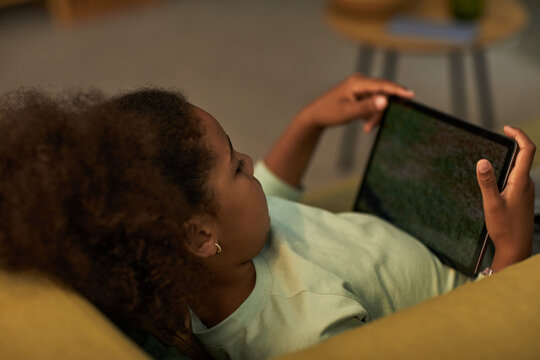 Black girl lying on sofa holding digital tablet with both hands watching screen, relaxing at home, curly hair visible, casual setting, technology use by child