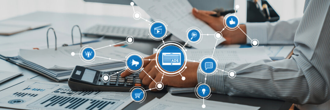 A business professional engaged in financial analysis using a calculator on a desk filled with documents and surrounded by digital business icons representing various themes and metrics. Amity