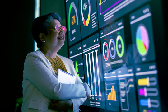 A senior asian businesswoman thoughtfully observes financial data displayed on a large screen monitor, contemplating market trends and strategic decisions.