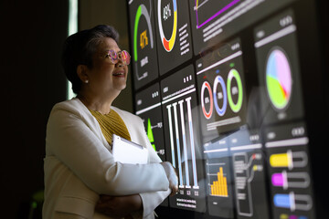 A senior asian businesswoman thoughtfully observes financial data displayed on a large screen monitor, contemplating market trends and strategic decisions.
