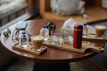 Red compact thermos, two glass tumblers with ice, and a glass of cappuccino arranged on a round wooden table