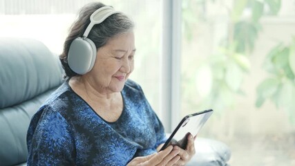 Asian senior woman sits comfortably on her sofa at home, eyes closed, happily listening to music on a digital tablet. - Powered by Adobe
