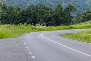 Winding asphalt road curves through the green summer landscape, leading toward the mountains under an expansive sky