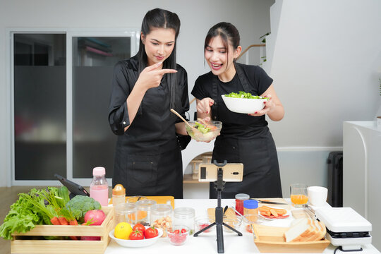Two asian women cooking food, making breakfast and salad while using smart phone or digital tablet, streaming live video clip or taking selfie photo together with happiness and fun in kitchen at home	