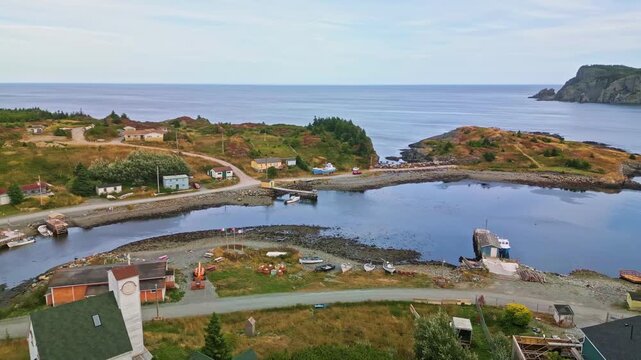 A drone flies over Brigus South harbour, capturing wooden wharves, fishing boats and colourful houses along the calm water and rocky coastline on Newfoundlands coast.