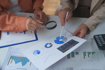 Two businesswomen collaborating on financial analysis, using a magnifying glass and pen to examine charts and graphs for informed decision-making on market trends