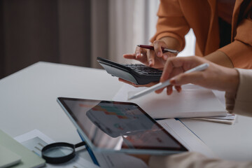 Businesswomen collaborating on financial analysis, using a calculator and tablet, pointing at graphs on the screen, surrounded by documents and a magnifying glass