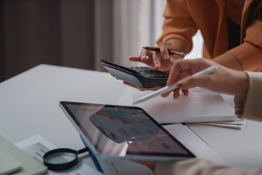 Businesswomen collaborating on financial analysis, using a calculator and tablet, pointing at graphs on the screen, surrounded by documents and a magnifying glass
