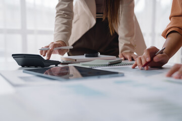 Two businesswomen are collaborating on a project, using a calculator and reviewing financial charts at an office desk, showcasing teamwork and financial analysis