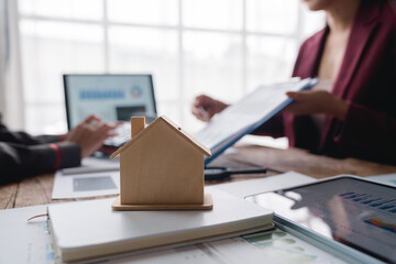 Real estate agent holding contract and explaining details to client during a meeting in the office, with wooden house model on the foreground