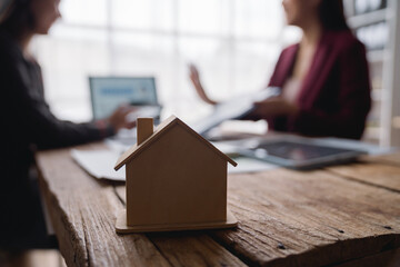 Small wooden house model sitting on a desk during a real estate meeting, with an agent and client discussing property investment and home ownership in the background
