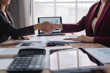 Two businesswomen shaking hands after reaching an agreement on a deal, sitting at a desk with financial documents, laptop, calculator, and tablet