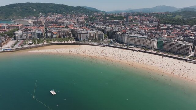 Aerial view of La Concha Bay in San Sebasti&aacute;n on a sunny summer day, with the beach full of people, turquoise water, moored boats, and the cityscape blending into the surrounding green hills