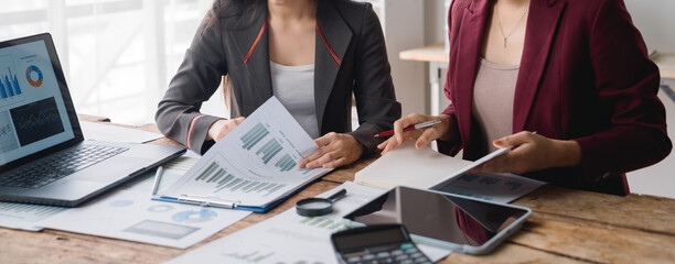 Two businesswomen collaborating at a desk, analyzing financial charts and graphs on a laptop,...